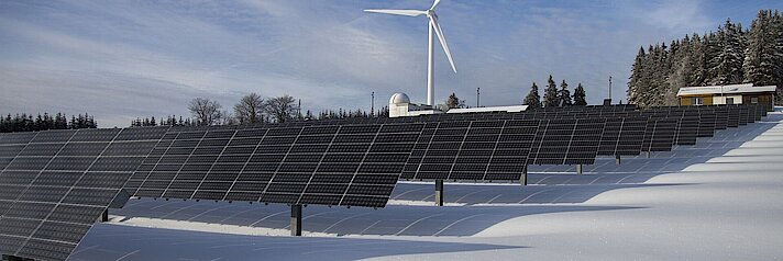 Im Vordergrund Solarpaneele, im Hintergrund ein Windrad inmitten einer Schneelandschaft