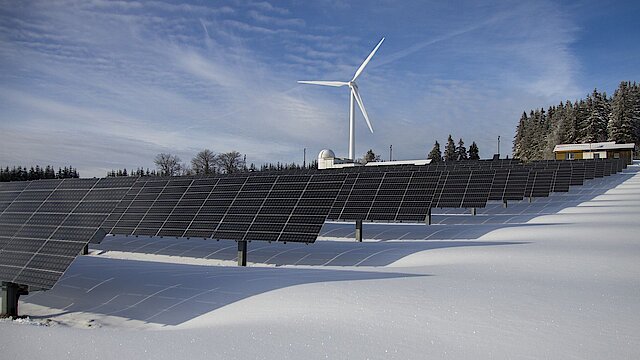 Im Vordergrund Solarpaneele, im Hintergrund ein Windrad inmitten einer Schneelandschaft